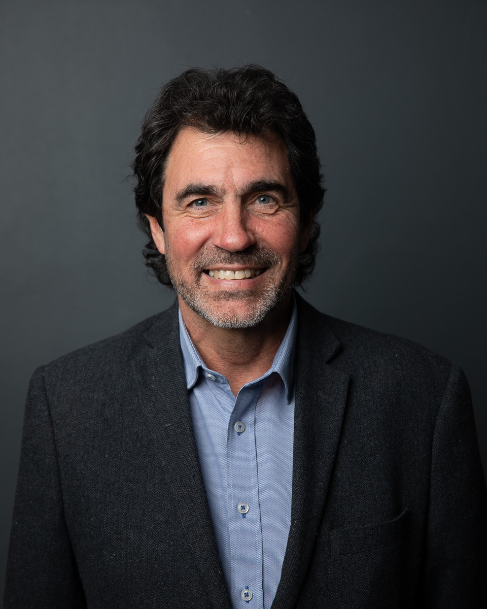 A corporate headshot of a smiling man in a blazer against a dark backdrop